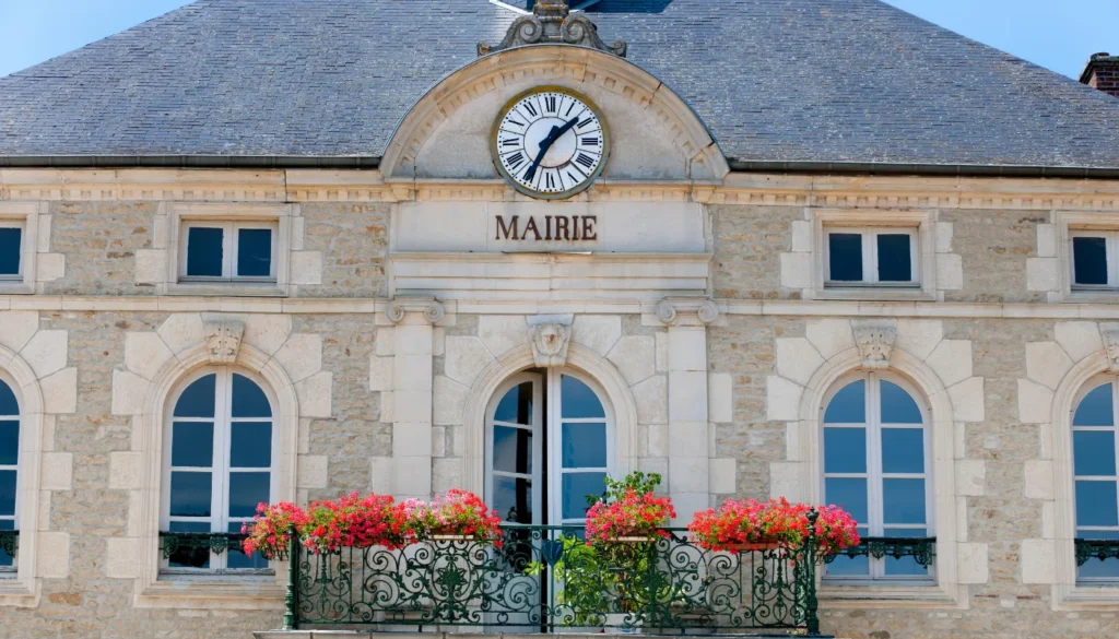 Bâtiment de la mairie avec horloge, fenêtres et balcon fleuri, architecture en pierre, sous un ciel bleu clair.